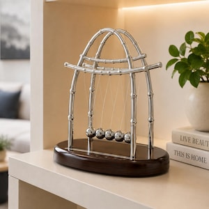 Puede incluir: Una cuna de Newton plateada con un marco diseñado para parecerse a tallos de bambú. El dispositivo consta de cinco esferas de metal pulido suspendidas por hilos finos de la estructura arqueada, montadas sobre una base de madera ovalada oscura.