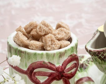 May include: A ceramic sugar bowl filled with brown sugar cubes. The bowl is light green with a dark red bow design. The bowl is on a matching tray with a creamer. White flowers are in the foreground.
