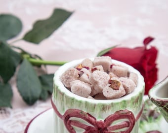 May include: A close-up of a teacup filled with sugar cubes, some with visible rose petals. The teacup has a green and white design with a red bow. A red rose and green leaves are in the background.
