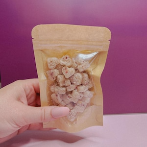 May include: A clear, resealable bag filled with heart-shaped candies. The candies are a light color with red flecks, and the bag is held against a pink and purple background.