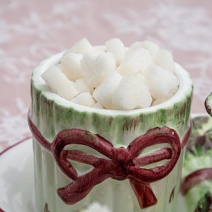 May include: A ceramic sugar bowl filled with white sugar cubes. The bowl is cylindrical with a green rim and a burgundy bow design. The bowl sits on a white saucer, with a blurred background.