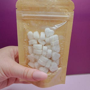 Puede incluir: Una bolsa de plástico transparente con un respaldo de papel marrón, llena de cubitos de azúcar blancos en forma de corazón. La bolsa se sostiene contra un fondo púrpura.