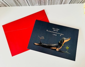 May include: A greeting card with a dachshund illustration and a red envelope. The card features a black and tan dachshund with a yellow collar, lying down next to a yellow flower. The card has text in German.