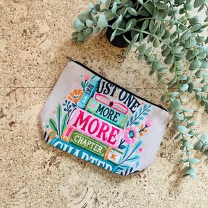 Op de afbeelding: Een lichtgrijze etui met ritssluiting met een kleurrijk ontwerp met gestapelde boeken en de tekst "Just One More Chapter". Het ontwerp bevat bloemen en botanische elementen in roze, blauwe en groene tinten.