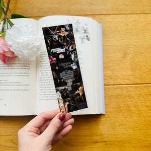 Può includere: Un segnalibro rettangolare con un design a collage con immagini e testo dello show "Vampire Diaries". Il segnalibro riporta le parole "Always Forever" e "Mystic Falls Virginia". Il segnalibro è tenuto sopra un libro aperto.