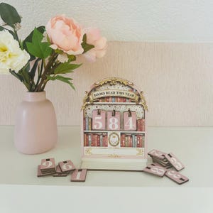 Peut inclure: Un calendrier en bois rose en forme d'étagère avec le texte "BOOKS READ THIS YEAR" au-dessus des chiffres 584. Des tuiles numérotées supplémentaires sont éparpillées. Un vase rose avec des fleurs est à gauche.
