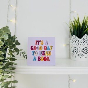 May include: A square, lilac sign with the text "IT'S A GOOD DAY TO READ A BOOK" in colourful, retro-style lettering. The sign is on a white shelf, with a potted plant to the left and a plant in a white, patterned pot to the right.