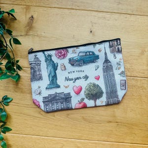 May include: A cream-colored zippered pouch featuring a New York City design. The design includes illustrations of the Statue of Liberty, the Empire State Building, a vintage car, and the text "New York New your city".