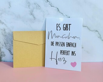 May include: White card with black text that reads "Es gibt Menschen, die passen einfach perfekt ins Herz" with a pink heart. The card is sitting on a brown envelope.