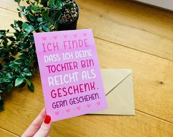 May include: A pink greeting card with the text "ICH FINDE, DASS ICH DEINE TOCHTER BIN REICHT ALS GESCHENK. GERN GESCHEHEN." is held in a hand. A brown envelope and a small potted plant are also visible.
