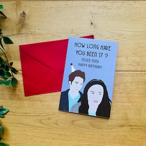 Könnte beinhalten: Eine Geburtstagskarte mit dem Text "HOW LONG HAVE YOU BEEN 17? NEVER MIND. HAPPY BIRTHDAY!" und Illustrationen von zwei Personen. Ein roter Umschlag liegt neben der Karte auf einer Holzoberfläche.