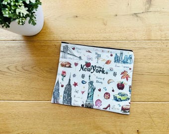 Op de afbeelding: Een witte etui met ritssluiting met een thema van New York City. Het ontwerp bevat illustraties van het Vrijheidsbeeld, het Empire State Building, een gele taxi en de Brooklyn Bridge. De woorden "New York City" zijn ook zichtbaar.