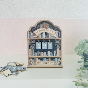 Op de afbeelding: Houten boekenplankkalender met de tekst "BOOKS READ THIS YEAR" en "MY LIBRARY". De kalender toont het nummer "197" en bevat kleine nummertegels. Het ontwerp bevat illustraties van boeken in blauw- en goudtinten.
