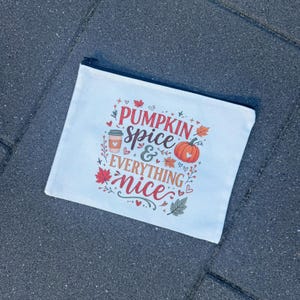 Könnte beinhalten: Eine weiße Reißverschlusstasche aus Segeltuch mit einer bunten herbstlichen Grafik und dem Text PUMPKIN SPICE & EVERYTHING nice. Das Design zeigt einen Kaffeebecher, einen orangefarbenen Kürbis und verschiedene Herbstblätter in Rot, Orange und Grün.