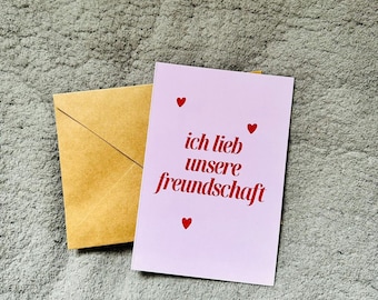May include: A light pink greeting card with the text "ich lieb unsere freundschaft" in red, accompanied by three red heart illustrations. A brown envelope is placed next to the card.