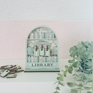 Könnte beinhalten: Hellblauer Holz-Ewiger Kalender im Bibliotheksstil mit dem Text "BOOKS READ THIS YEAR" und "LIBRARY". Der Kalender zeigt die Zahlen "112". Zusätzliche Zahlenkacheln sind in der Nähe verstreut. Eine dekorative Pflanze befindet sich im Hintergrund.