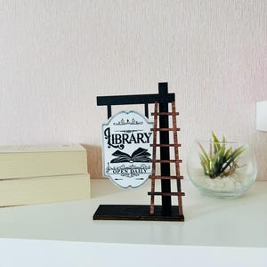 May include: A black and brown wooden sign featuring the word "LIBRARY" and an open book graphic. The sign has a small ladder and is on a black base. The sign reads "OPEN DAILY".