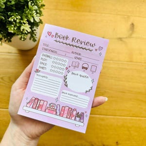 Könnte beinhalten: Ein helllila Notizblock für Buchrezensionen mit dem Wort "Book Review" oben. Der Notizblock hat Platz für Titel, Autor und Genre, mit Bewertungssternen und Abschnitten für Gedanken, Lieblingszitat und Moment. Unten ist eine kleine Bücherregal-Illustration.