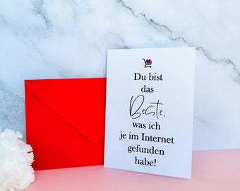 May include: A white greeting card with black text and a red shopping cart icon, next to a bright red envelope. The card reads "Du bist das Beste, was ich je im Internet gefunden habe!"