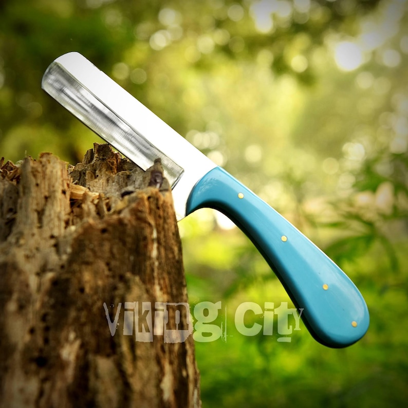 May include: A straight razor with a silver blade and a light blue handle with gold-colored rivets. The razor is resting on a weathered tree stump. The text "Viking City" is visible on the stump.
