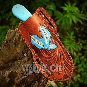 May include: A brown leather knife sheath with a light blue knife handle. The sheath features three blue and white feather designs and the text "Viking City". The sheath is made of brown leather and is designed to hold a knife.