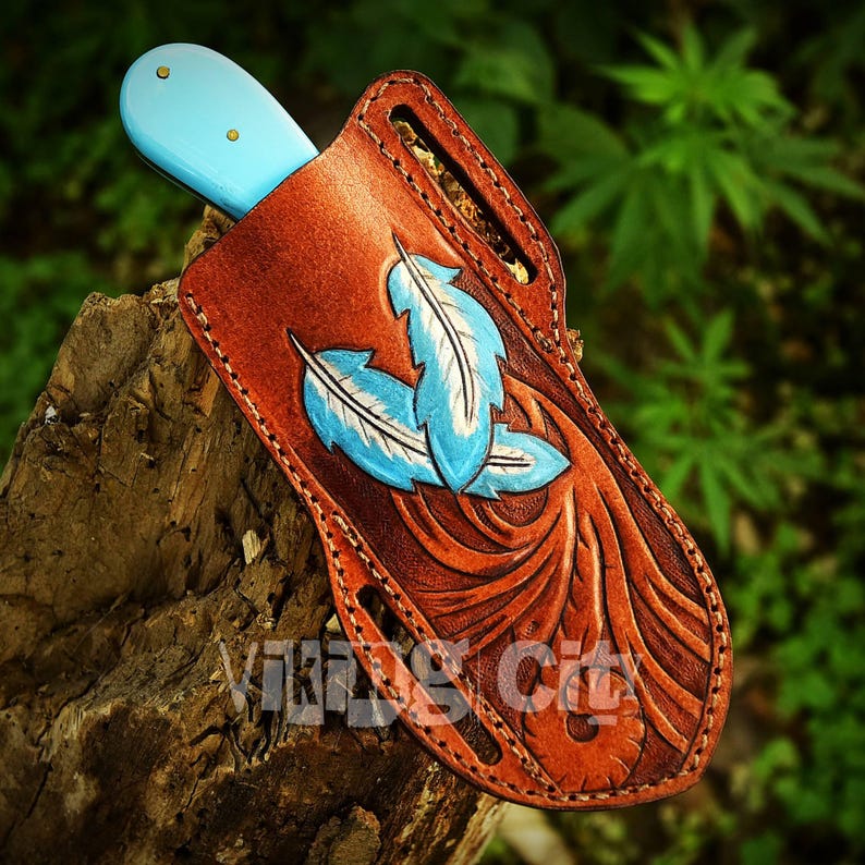 May include: A brown leather knife sheath with a light blue knife handle. The sheath features three blue and white feather designs and the text "Viking City". The sheath is made of brown leather and is designed to hold a knife.