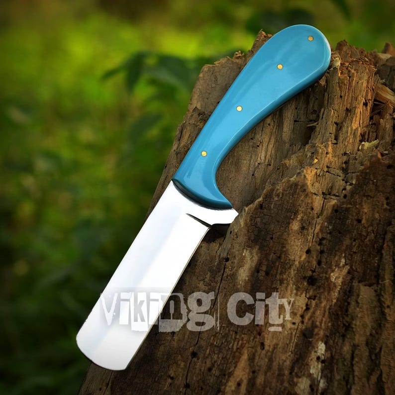 May include: A knife with a bright blue handle and a polished silver blade. The handle is secured with three gold-colored rivets. The text "Viking City" is printed on the wooden surface. The knife is displayed against a blurred green background.