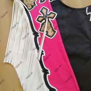 Op de afbeelding: Een close-up van een leren chaps. De chaps hebben een zwart en roze ontwerp met een kruis en bloemaccenten. Witte franjes sieren de zijkant. De chaps hebben een bruine riem en zijn waarschijnlijk voor paardrijden.