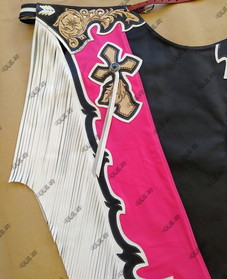 Op de afbeelding: Een close-up van een leren chaps. De chaps hebben een zwart en roze ontwerp met een kruis en bloemaccenten. Witte franjes sieren de zijkant. De chaps hebben een bruine riem en zijn waarschijnlijk voor paardrijden.