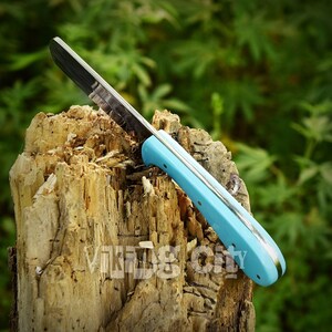 May include: A knife with a black blade and a light blue handle. The handle has a silver trim and is attached to the blade with rivets. The knife is resting on a weathered wooden stump. The text "Vikings City" is visible on the stump.