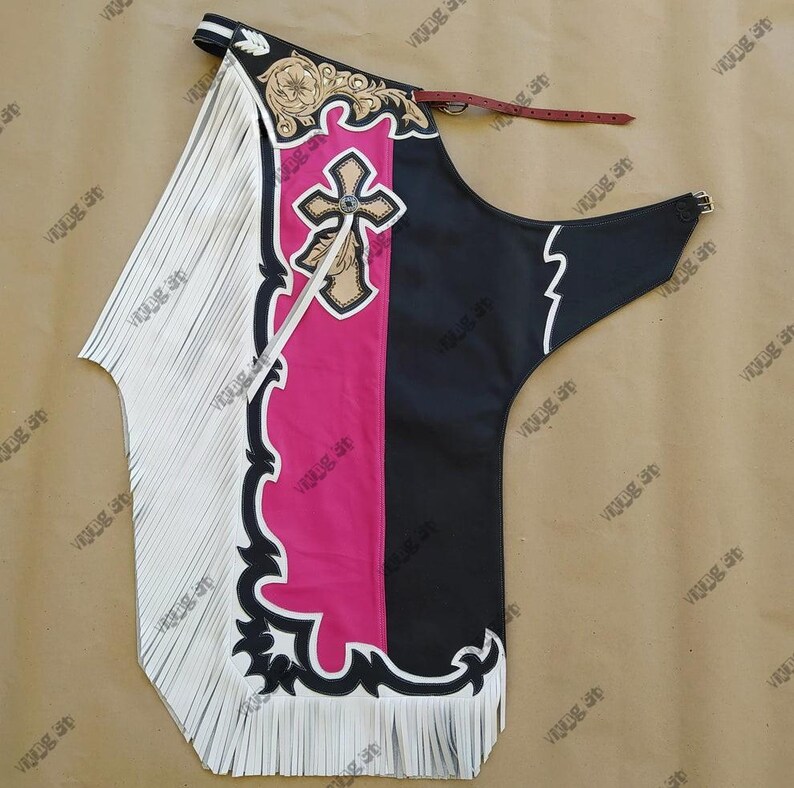Op de afbeelding: Westerse chaps met een wit gefranjerd paneel, een roze middenpaneel en een zwart paneel. De chaps hebben een kruisontwerp, decoratieve stiksels en een leren riem. Ontworpen om te paardrijden.