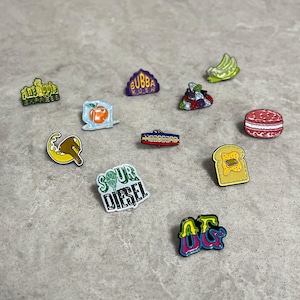 Könnte beinhalten: Eine Sammlung von elf bunten Emaille-Anstecknadeln mit verschiedenen Motiven, darunter Schriftzüge wie Pineapple Express, Bubba Kush, Headband und Sour Diesel, sowie Symbole von Früchten, einem Macaron, einer Scheibe Toast und abstrakten Formen.