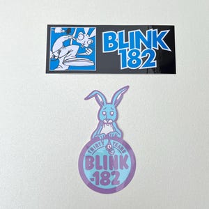 Pode incluir: Dois adesivos, um retangular e outro circular, com o logotipo Blink-182. O adesivo superior é preto com um gráfico de coelho azul e o texto BLINK 182. O adesivo inferior mostra um coelho azul claro com um relógio de bolso sobre o texto THIRTY YEARS BLINK-182.