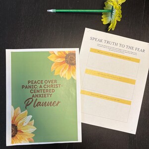 May include: A planner titled "Peace Over Panic: A Christ-Centered Anxiety Planner" with a green cover and sunflower illustrations. A pen with a yellow flower is on top of the planner and a worksheet that says "Speak Truth to the Fear".
