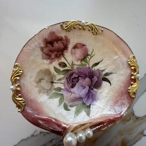 May include: A decorative, shell-shaped dish with a floral design. The dish features a painted bouquet of purple and pink peonies on a cream-colored background. Gold filigree accents and faux pearls adorn the edges, creating an elegant, vintage aesthetic.