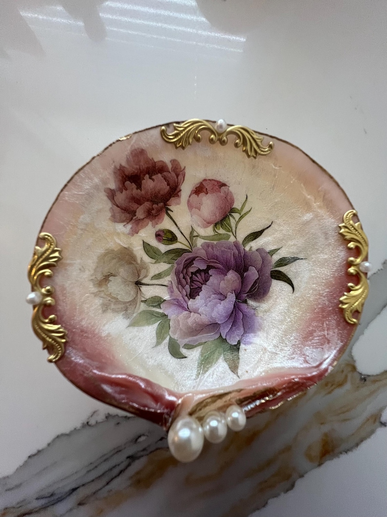 May include: A decorative, shell-shaped dish with a floral design. The dish features a painted bouquet of purple and pink peonies on a cream-colored background. Gold filigree accents and faux pearls adorn the edges, creating an elegant, vintage aesthetic.