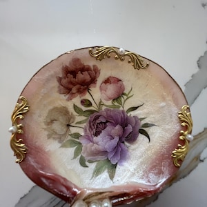 May include: A decorative shell-shaped dish with a floral design. The dish features a bouquet of purple and pink peonies with green leaves. The edges are trimmed with gold accents and faux pearls. The shell has a pink and cream color scheme.