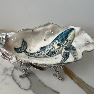 Puede incluir: Un plato decorativo de concha de ostra con un diseño de ballena azul pintado a mano. La ballena presenta patrones florales y se encuentra en un interior nacarado. La concha está sostenida por una base de metal plateado y adornada con pequeños cristales.
