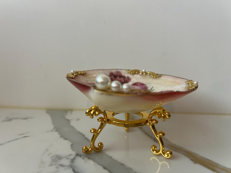 May include: A decorative shell-shaped dish with a floral design and pearl accents, resting on a gold-colored, ornate stand. The dish features a cream-colored interior with red and pink floral details and is trimmed with gold.