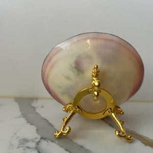 May include: A decorative seashell, cream-colored with pink edges, is displayed on a gold-colored metal stand. The stand has a circular top and ornate, curved legs, adding an elegant touch to the presentation.
