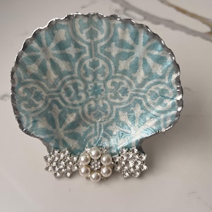 May include: A decorative seashell-shaped trinket dish. The dish features a light blue and white patterned interior with a silver-colored edge. The base is adorned with pearl and rhinestone embellishments. Ideal for holding jewelry or small items.