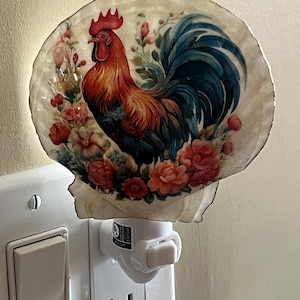 Puede incluir: Luz de noche decorativa con un diseño de gallo colorido. El gallo está representado con tonos rojos, naranjas y azules, rodeado de acentos florales rosas y rojos. La luz de noche se enchufa a un enchufe de pared estándar.