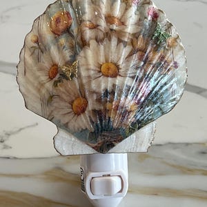 Könnte beinhalten: Dekoratives Nachtlicht mit Muscheldesign. Die Muschel ist mit einem Blumenarrangement aus Gänseblümchen und Rosen verziert, mit einem goldenen Schmetterlingsakzent. Das Nachtlicht hat einen weißen Sockel mit einem Ein-/Ausschalter.