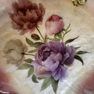 May include: Decorative, shell-shaped tray with a floral design. The tray features a bouquet of peonies in shades of purple, pink, and burgundy, with green leaves. The rim is a light pink color, and the tray has gold accents.