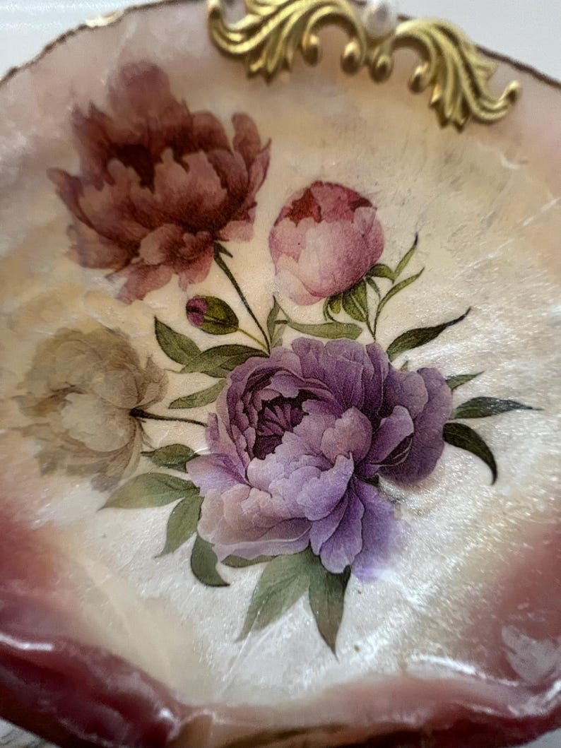 May include: Decorative, shell-shaped tray with a floral design. The tray features a bouquet of peonies in shades of purple, pink, and burgundy, with green leaves. The rim is a light pink color, and the tray has gold accents.