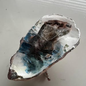 May include: A decorative oyster shell-shaped dish featuring a detailed image of an otter in water. The dish has a silver-colored trim and a small metal stand. The otter is brown and white, and the water is depicted in shades of blue.