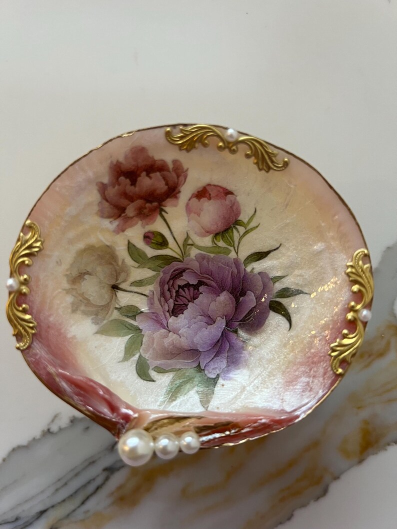 May include: A decorative seashell-shaped dish with a floral design. The dish features a pink and gold rim, with a painted bouquet of purple and red peonies. Gold filigree and pearls adorn the edges, creating an elegant, feminine aesthetic.