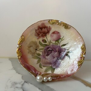 May include: A decorative seashell dish with a floral design featuring pink and purple peonies. The dish has gold-colored accents and faux pearls along the edge. The shell is approximately 4 inches in diameter.