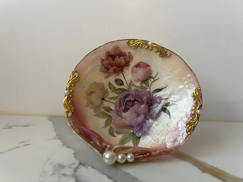 May include: A decorative seashell dish with a floral design featuring pink and purple peonies. The dish has gold-colored accents and faux pearls along the edge. The shell is approximately 4 inches in diameter.