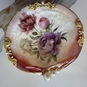 May include: A decorative, shell-shaped dish with a floral design. The dish features a painted arrangement of purple, pink, and white peonies with green leaves. Gold filigree accents the edges, and small pearls are placed around the rim.